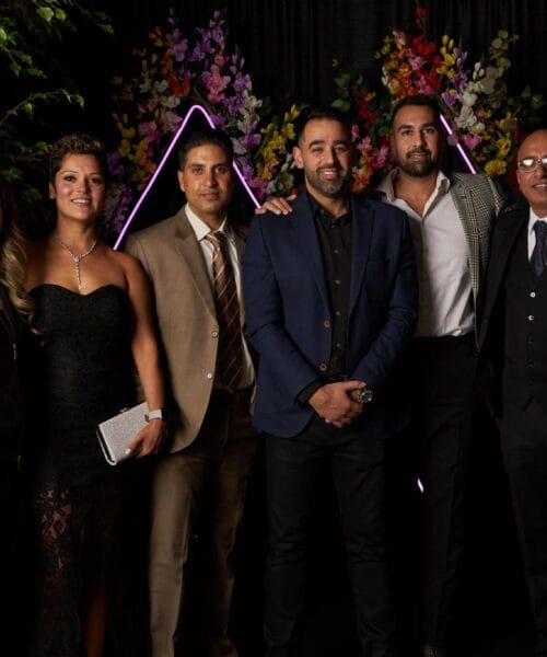 A group of six people dressed in semi-formal to formal attire pose together in front of a backdrop decorated with greenery and colorful flowers. They are smiling and appear to be at an event or celebration.