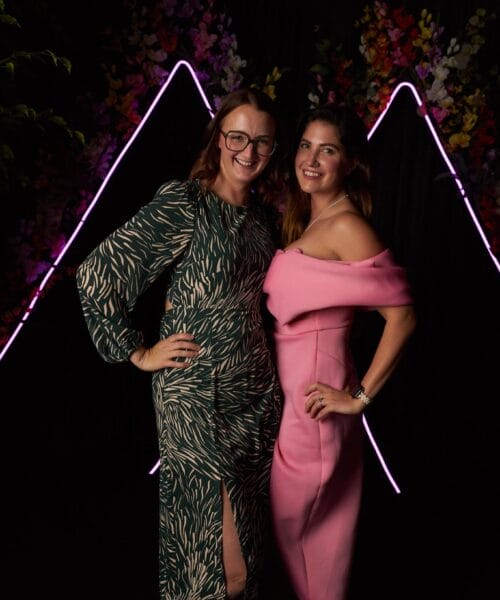 Two women are smiling and posing together against a dark backdrop with neon lights and floral decorations. The woman on the left is wearing glasses and a green patterned dress, while the woman on the right is dressed in a pink off-the-shoulder dress. The lighting is low.