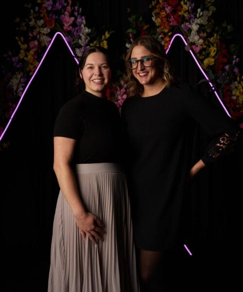 Two women are posing and smiling in front of a dark backdrop with purple neon lights. One woman is wearing a black top and a grey skirt, and the other is in a black dress. Colorful flowers and green plants are in the background, adding a vibrant touch.