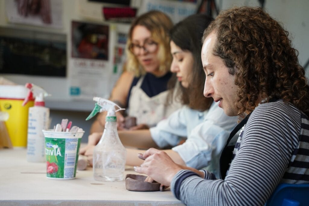 Three people sit at a table shaping clay with their hands, focused on their pottery work. Art supplies and water spray bottles are on the table. The scene is set in a creative, casual studio environment.