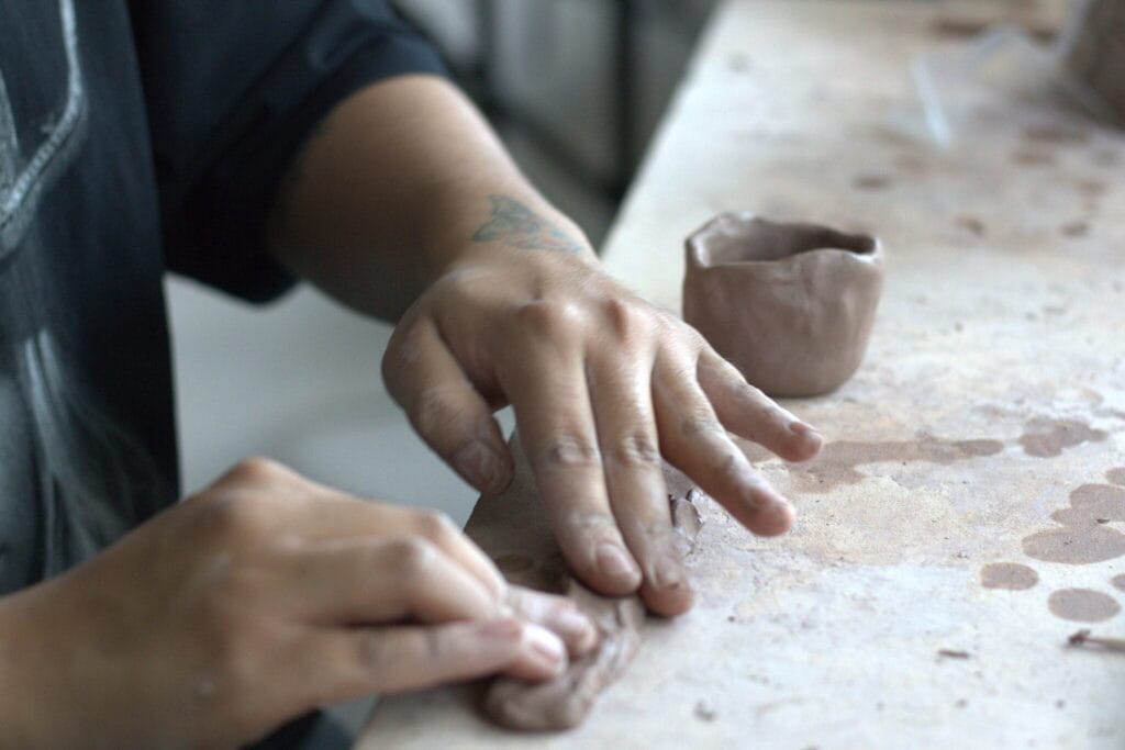 A person shapes clay with their hands on a worktable. A small, unfinished clay pot sits nearby, and the person’s fingers are coated in clay.