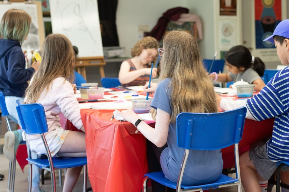Un groupe d'enfants est assis autour d'une table recouverte d'une nappe rouge et peint sur du papier. Des fournitures artistiques et des pots de peinture sont posés sur la table. Les enfants semblent concentrés et absorbés par leur activité.