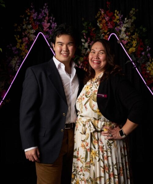 A young man in a blue blazer and a woman in a floral dress stand side by side, smiling, in front of a backdrop adorned with colorful flowers and purple neon lights forming triangular shapes. Green foliage frames the edges of the image.