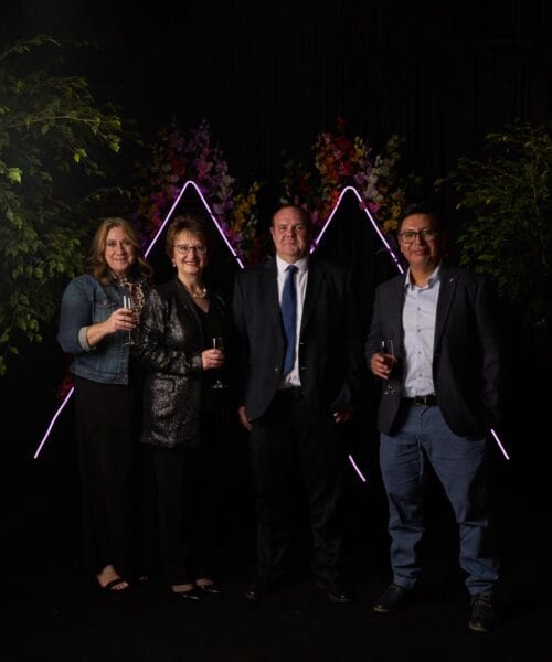 Four people dressed in formal attire stand in front of a backdrop with neon triangular lights and floral decorations. Two women are on the left, holding wine glasses, and two men are on the right, smiling. The scene appears to be at a celebratory event.