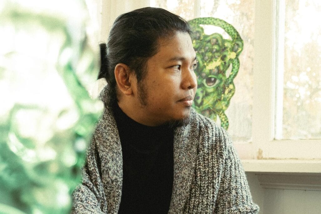 A person sitting in a room with natural light, wearing a gray cardigan and black top. They have long hair tied back and appear to be looking out of the window. A green glass sculpture is in the foreground.