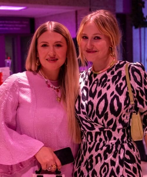 Two women are posing and smiling in a well-lit indoor setting with a purple hue. The woman on the left is wearing a light pink dress, holding a phone. The woman on the right is wearing a black and white animal print dress, holding a drink. The background has some signs and a bar.