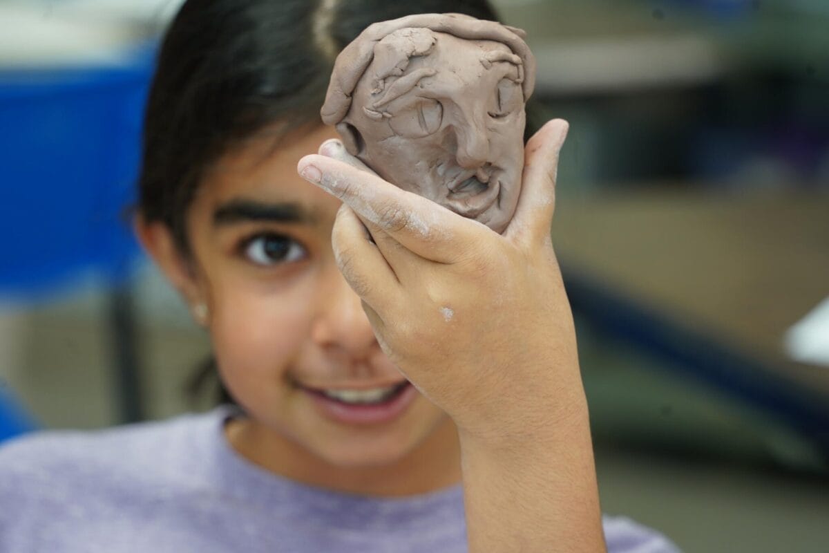 Un enfant aux cheveux noirs, vêtu d'une chemise violette, tient une petite sculpture en argile représentant un visage, occultant partiellement son propre visage. La sculpture semble être en cours de réalisation avec des détails visibles comme des yeux, un nez et une bouche.
