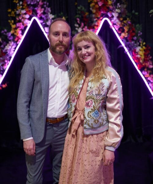 A man and a woman stand together, smiling at the camera. They are dressed formally, with the man in a gray suit and the woman in a pink dress and patterned jacket. Behind them are illuminated, purple triangular lights and floral decorations, set against a dark background.