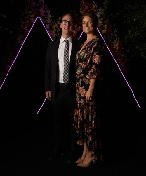 A man and woman dressed formally stand in front of a dark backdrop with neon pink triangular light designs and colorful floral decorations. The man is wearing a suit, tie, and glasses, while the woman is in a floral dress and beige heels. Both are smiling.