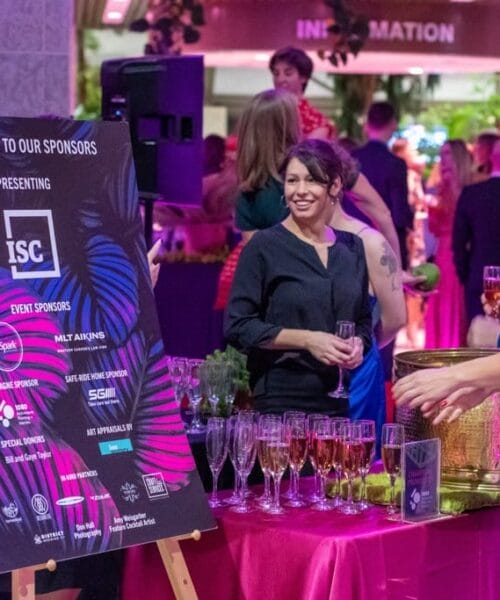 A group of people socializes at an indoor event. A woman wearing a black dress stands behind a table filled with glasses of champagne. Nearby, an easel displays a sign thanking event sponsors. The venue is decorated with plants and has a festive atmosphere.