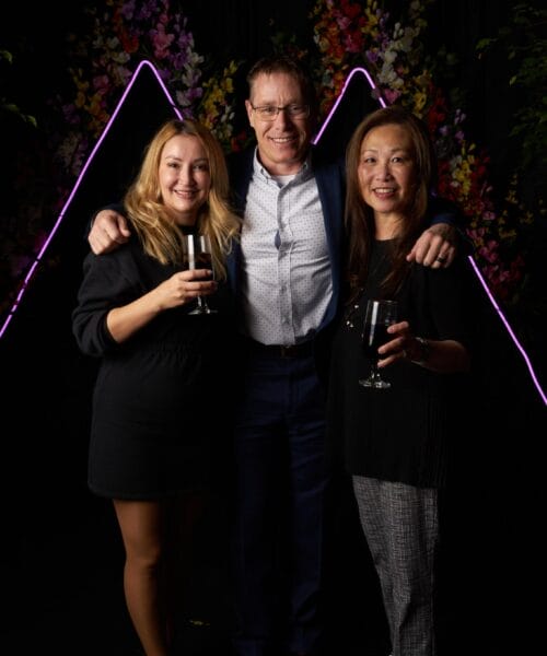 Three people stand together, smiling and holding drinks. The person on the left is wearing a black dress, the person in the middle is wearing a light blue shirt and a dark jacket, and the person on the right is wearing a black blouse and checkered pants. There is a colorful backdrop.