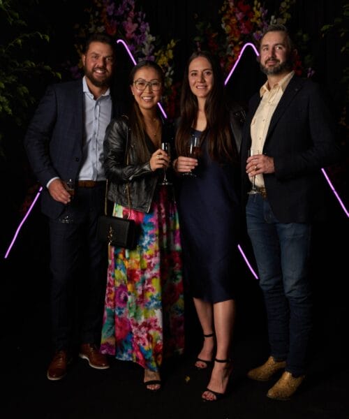 Four people stand together, smiling and holding drinks in front of a backdrop with purple neon lights and floral decorations. Two men in suits and two women in dresses and jackets are posing; one woman is wearing a colorful patterned dress, and the other a navy blue dress.