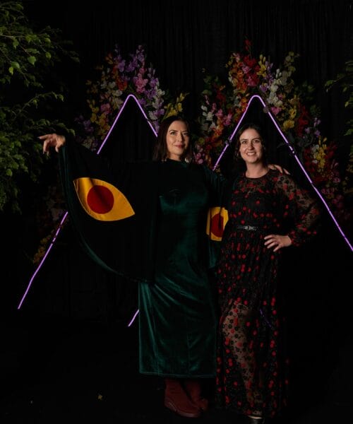 Two people stand in front of a dark backdrop with vibrant floral arrangements. One wears a green dress with bold eye designs on the extended sleeves, resembling wings. The other wears a black and red dress, smiling with arm around the first person's waist. Neon triangles frame them.