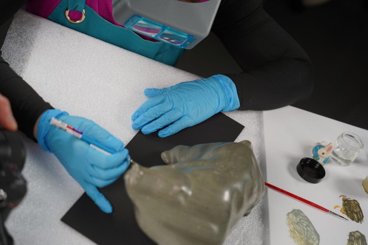 A person wearing blue gloves and a magnifying headset works on a small sculpture. They hold a precision tool, focusing on details. Nearby are paintbrushes, a camera, and small containers on the table.