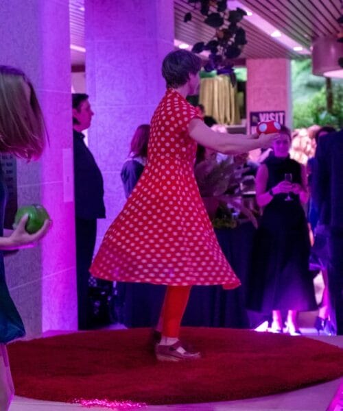 A person in a red polka dot dress is performing a dance on a red circular platform under purple lighting. Onlookers in formal attire are watching in a stylish indoor setting.