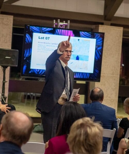 An auctioneer stands at the front of a room, pointing upwards with his left hand while holding a sheet of paper in his right. Behind him, a display screen shows details about 
