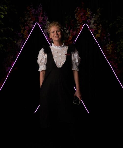 A person with short, curly blonde hair stands smiling in front of a backdrop of illuminated pink and purple triangular neon lights. They are wearing a black overall dress with a white puff-sleeve blouse featuring a textured dot pattern. Trees and flowers frame the scene.