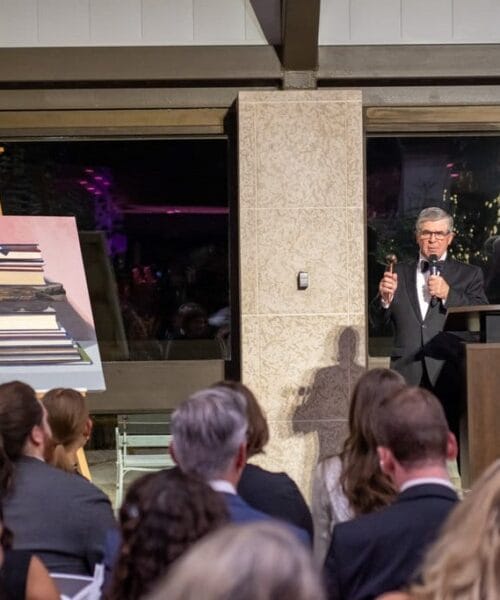 A man in a suit and tie stands at a podium, speaking into a microphone at an indoor event. Behind him is a large window showing a night scene and a painting of stacked books. Seated audience members face the speaker, listening attentively.