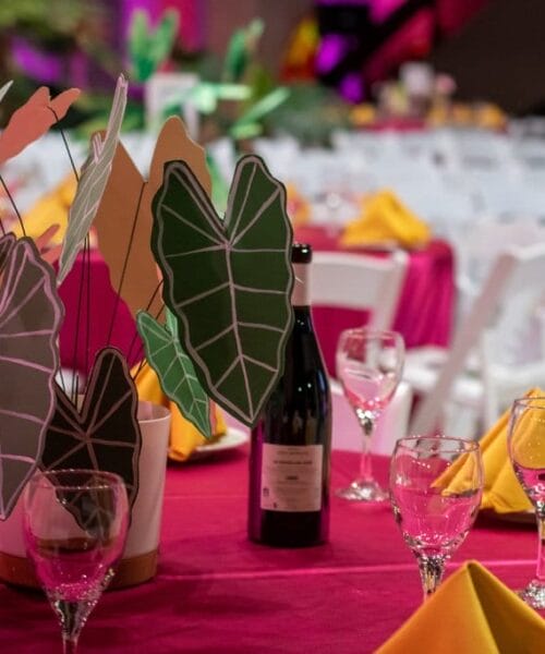 A table set for a banquet with red tablecloths, yellow napkins, and clear wine glasses. A bottle of wine and a centerpiece featuring green, pink, and white decorative leaves are on the table. More tables are visible in the background.
