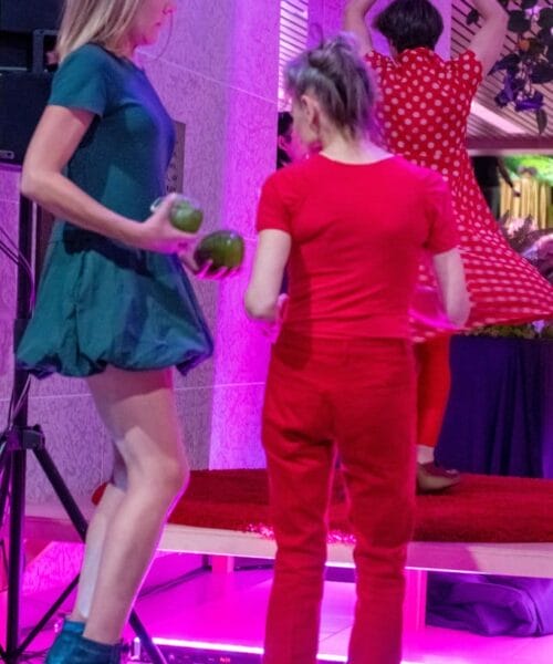 Three women are performing on a stage. The woman on the left is juggling green clubs. The woman in the middle, standing on the floor in front of the stage, is dressed in red. The woman on the right, on stage, is spinning, wearing a red polka dot dress and matching red tights.