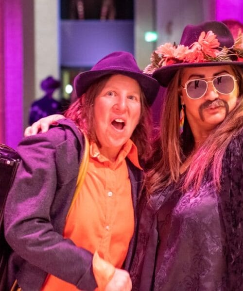 Two people in festive costumes pose for a photo at a vibrant indoor event. One wears a black hat, orange shirt, and has an accordion. The other dons a flower crown, black hat, large sunglasses, and a fake mustache. The backdrop is lit with colorful pink lighting.