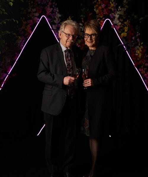 An elderly man and woman stand together, both dressed formally, holding glasses of champagne. They smile at the camera, with neon-lit triangular shapes and colorful flowers forming a visually striking backdrop.