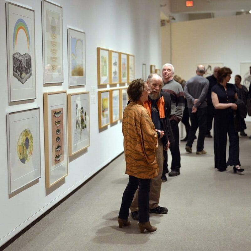 Les gens marchent et discutent dans une galerie d’art, regardant diverses œuvres d’art encadrées sur les murs. La galerie est bien éclairée et aux murs blancs, créant une atmosphère lumineuse et spacieuse. Les peintures et photographies sont exposées en couleur et en noir et blanc.