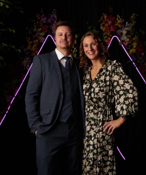 A man and a woman are posing together in front of a dark backdrop with neon-lit mountain shapes and floral decorations. The man is wearing a blue suit and tie, while the woman is dressed in a black and white floral dress, smiling with her hand on her hip.