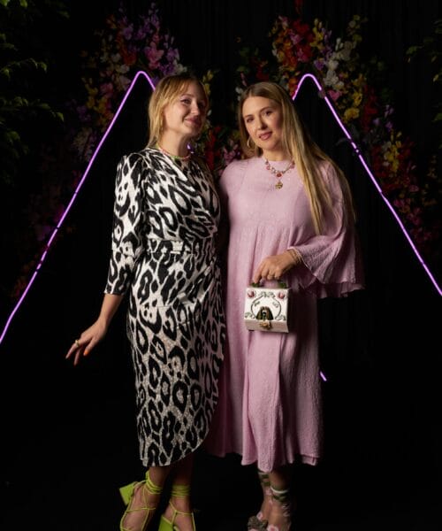 Two women are standing side by side in front of a backdrop with colorful flowers and neon triangular lights. The woman on the left is wearing a black and white animal print dress with green shoes, and the woman on the right is wearing a pink dress and holding a bag.