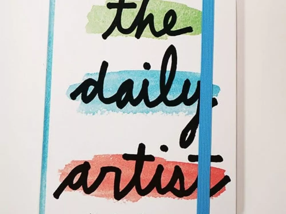 The image shows a journal with a white cover titled “the daily artist” in black, stylized handwriting. The words 