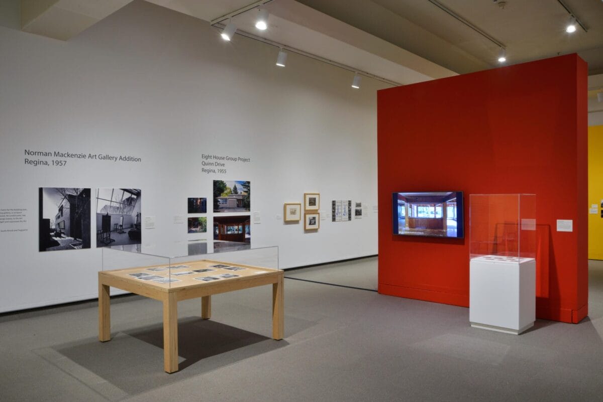 A modern art gallery interior featuring a display case with documents on a wooden table, framed artworks on white walls, and a prominent red wall with a small screen. Track lighting illuminates the exhibits, evoking the vibrant energy of Spring on the Prairie.