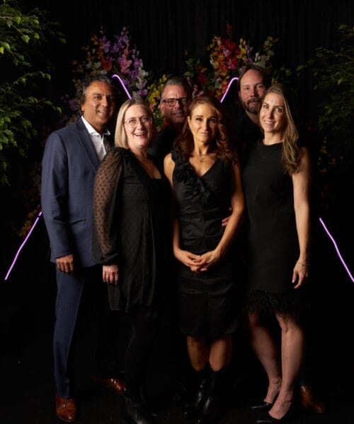 Six people are standing and posing for a group photo in front of a dark backdrop decorated with colorful flowers and purple lights. Three men and three women are dressed in formal attire, with the men in suits and the women in black dresses.