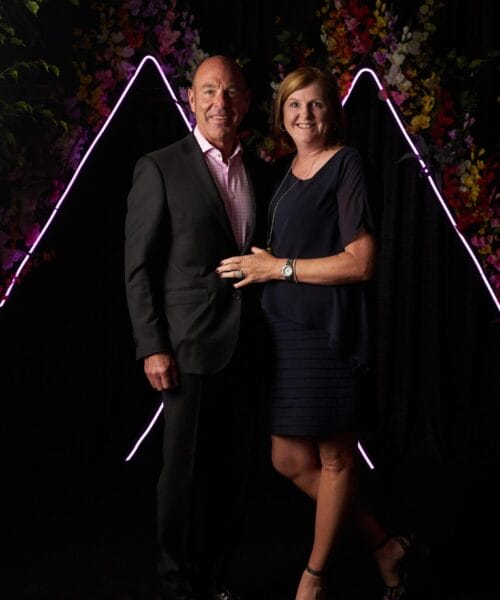 A man in a dark suit and a woman in a navy blue dress stand closely together, smiling at the camera. They are against a black backdrop with colorful flowers and neon pink lights forming a geometric pattern. The woman is wearing black heeled sandals.