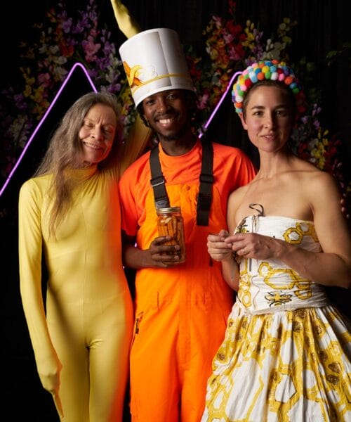 Three people in colorful costumes stand together against a dark background with floral decorations. One wears a yellow bodysuit, another wears an orange outfit with a tall hat, and the third wears a patterned dress with a pom-pom headband. They are all smiling.