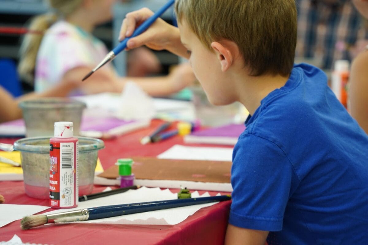 Un jeune garçon en chemise bleue peint sur une table couverte de matériel artistique. Des pinceaux, des pots d'eau et des bouteilles de peinture sont posés sur la table. D'autres enfants sont visibles à l'arrière-plan, également occupés à peindre.