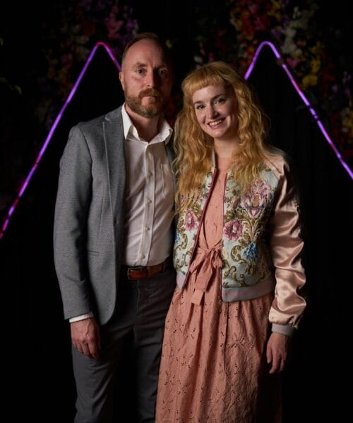 A man and a woman standing side by side, smiling at the camera. The man is wearing a gray suit with a white shirt, and the woman is wearing a pink dress with a floral embroidered jacket. The background is dark with colorful, illuminated triangular shapes and foliage.