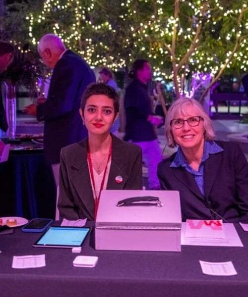 Two women sit at a table covered with a black tablecloth beneath strings of lights. On the table are a tablet, a cash box, signs and some paperwork. Both women are smiling at the camera. Other people are visible in the background, involved in various activities.