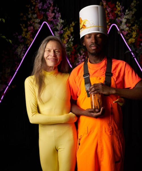 Two people stand in front of a floral backdrop with neon lights. The person on the left wears a yellow bodysuit and has long hair. The person on the right wears an orange outfit with suspenders and a white hat, holding a small trophy or award.