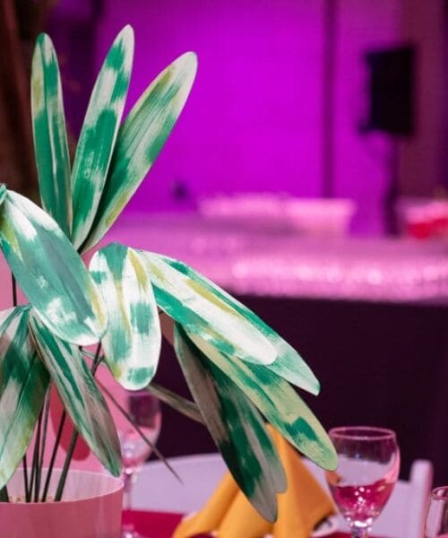 A decorative plant with green and yellow leaves is placed in a white pot on a table set for an event. The table also has wine glasses and yellow napkins. In the background, there are empty glasses arranged on a counter, with purple lighting in the room.
