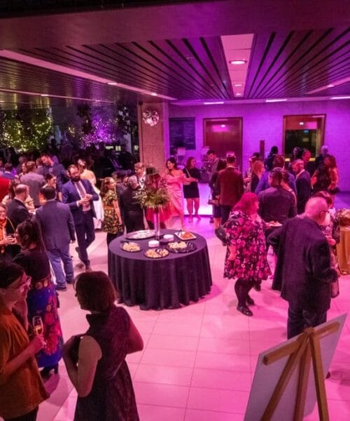 A crowded indoor event with people mingling and chatting in a room illuminated by pink and purple lighting. Several standing tables with snacks are scattered around, and a few attendees are dressed in formal attire. Green plants and decorations are visible in the background.