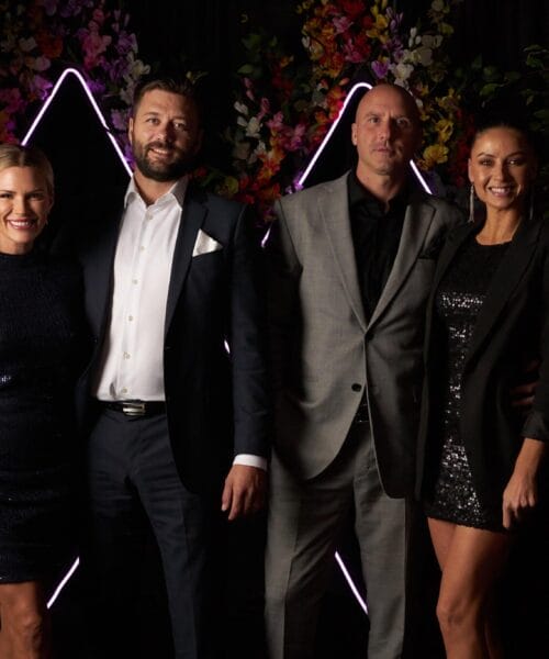 A group of four people dressed in formal attire stands in front of a colorful floral background with neon triangle lights. The group includes two women in sequined dresses and two men in suits, all smiling at the camera.