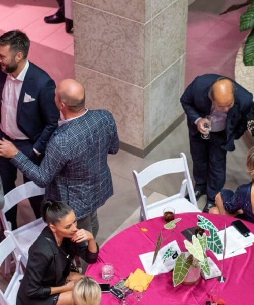 A group of people are gathered at a social event. Some are standing and talking while others are sitting at a table with a pink tablecloth and green leaf decorations. The atmosphere appears lively, and the setting includes plants and modern decor.