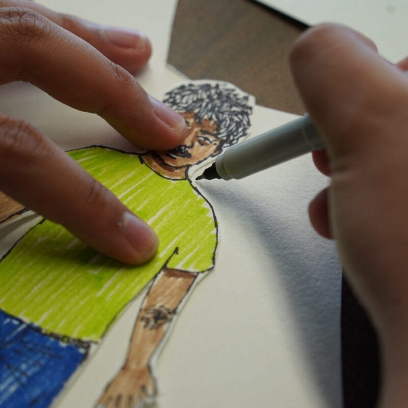 A person uses a black pen to outline the shoulder of a hand-drawn paper cutout figure wearing a green shirt and blue pants, while another hand holds the paper cutout in place.