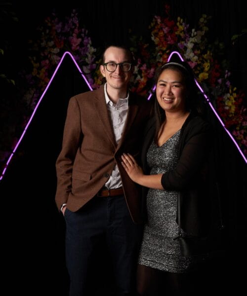 A man and a woman are dressed formally, standing in front of a black background adorned with floral decorations and neon light accents. The man is wearing a brown blazer, and the woman is in a sparkly dress with a black jacket, smiling with her arm around him.