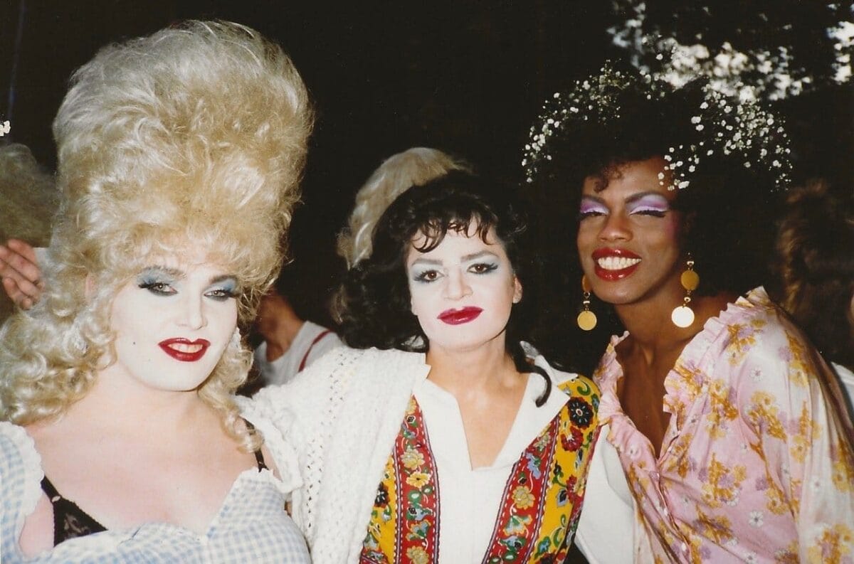 Three individuals pose for a photo. The person on the left has large, teased blonde hair, heavy makeup, and is wearing a gingham top. The middle person has short dark hair, white face makeup, and a colorful vest. The right person has curly hair with flowers and is wearing vibrant makeup and a floral top.