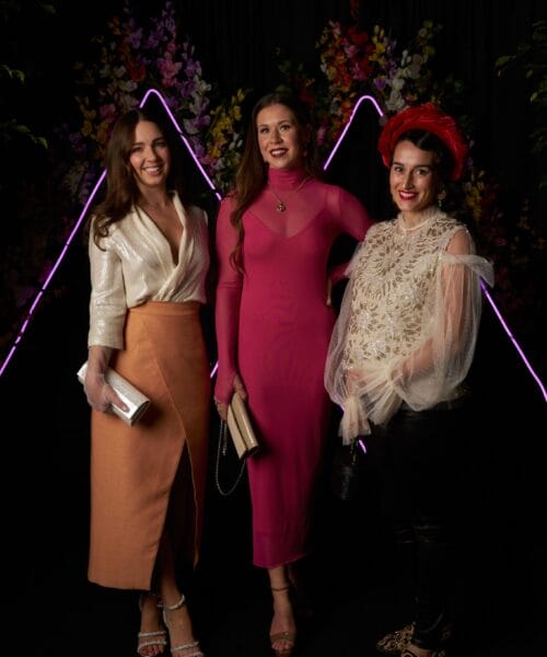 Three women pose together in elegant attire against a dark backdrop with decorative purple lighting. The left woman wears a white blouse and tan skirt, the middle woman wears a long pink dress, and the right woman wears a ruffled beige top and black pants with a red hat.