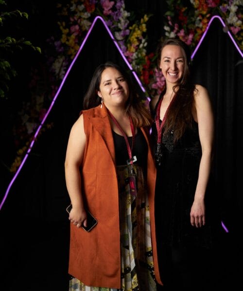 Two people stand smiling in front of a dark backdrop adorned with colorful flowers and purple neon triangle lights. The person on the left wears a brown sleeveless jacket and floral skirt, holding a phone. The person on the right wears a black dress with long hair down.