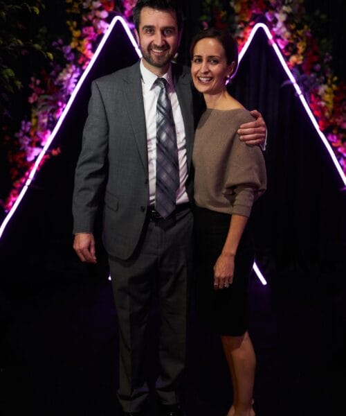 A man in a grey suit and tie stands beside a woman in a beige top and black skirt. They are smiling and posing in front of a backdrop decorated with colorful flowers and illuminated neon triangular shapes. The man has his arm around the woman's shoulder.