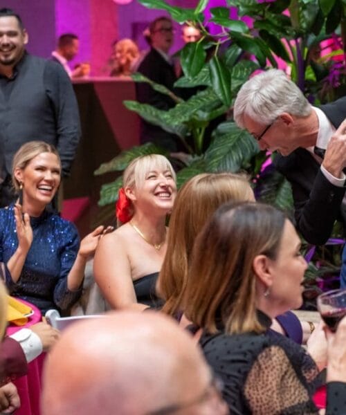 A group of people at a lively social gathering. They are seated around a table, laughing and chatting. One person in a black suit is standing, engaging with the others. Lush green plants and vibrant lighting create a festive atmosphere.