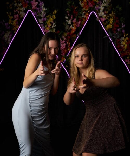 Two women strike playful poses in front of a black backdrop adorned with colorful flowers and neon purple triangular lights. One wears a light blue dress and the other a dark brown dress. They point finger guns towards the camera while smiling and crouching slightly.